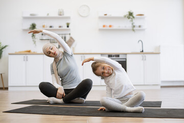 Slim muslim lady on yoga mat stretching arms while training with small female kid. Young parent and daughter in sportswear spend time happily during workout.