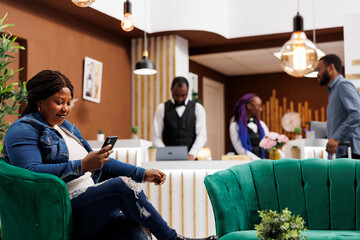Happy relaxed African American woman relaxing in hotel lobby using smartphone watching online video or browsing internet. Female tourist resting with phone in lounge area at luxury resort - obrazy, fototapety, plakaty