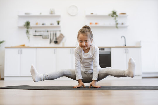 Preschool cute little girl practicing yoga, standing in crane exercise, bakasana pose, working out on mat wearing sportswear, indoor full length, in white loft kitchen background.