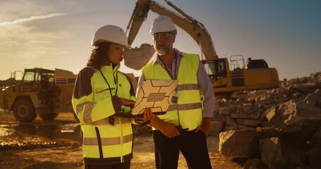 Caucasian Male Engineer And Hispanic Female Building Architect Walking On Construction Site With Laptop Computer And Talking About New Real Estate Project. Heavy Machinery Working. Golden Hour