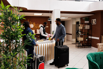 Happy black couple standing at hotel front desk with luggage, checking in at honeymoon resort,...