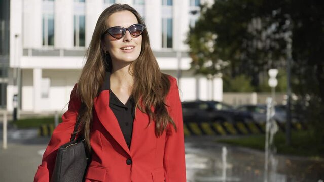 A young beautiful Caucasian businesswoman in sunglasses walks with a confident smile - a fountain and an office building in the blurry background - ends in closeup
