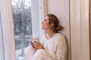 Close-up portraits of young stylish woman sitting on windowsill, drinking cocoa from cup, looking out window. Winter festive atmosphere. Girl in home clothes drinks tea, coffee. Holiday light bulbs.