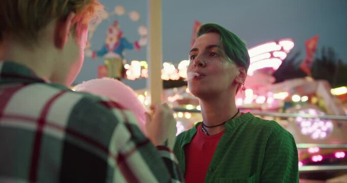 A blonde girl with a short haircut in a plaid shirt feeds her girlfriend with a short green haircut in a Green shirt and also passes a piece of pink cotton candy with her mouth during their date in a
