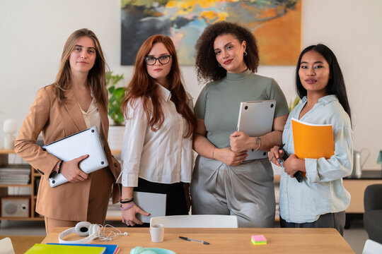 Group of cheerful young smiling women colleagues in smart elegant clothes standing near wooden table and looking at camera while holding laptops and notebook at office