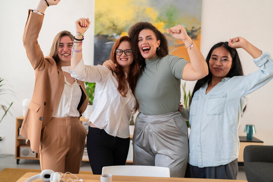 Happy young women colleagues raising arms and celebrating success while standing against blurred background in modern creative office and looking at camera