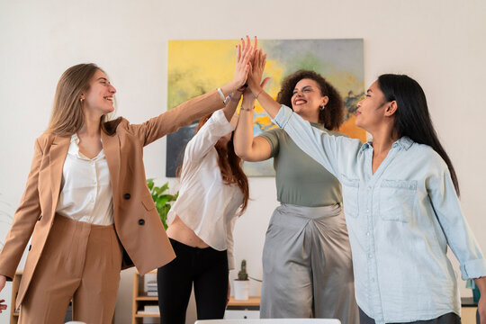 Happy young women colleagues standing and giving high five to each other while celebrating success in modern creative office during business meeting