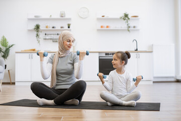 Athletic muslim woman and little girl lifting dumbbells while looking at each other on kitchen floor. Caring mother in activewear teaching inquisitive daughter strength exercises in apartment.