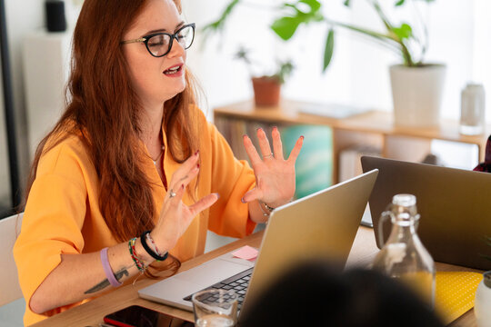 Thoughtful young female worker in casual clothes with makeup and eyeglasses sitting at table with laptops and showing hand gesture while talking and working on remote project in office