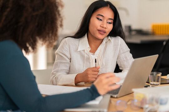 Positive young Asian female in casual clothes sitting at table with laptop while talking with colleague and taking notes in notebook during work hour on project in office
