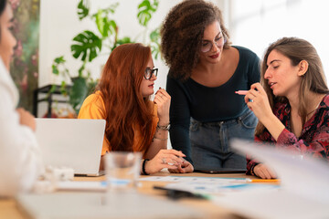 Positive young female coworkers in casual clothes gathered at wooden table studying and analyzing charts while working together on project in modern workspace