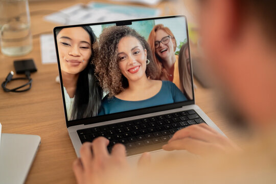 Laptop screen image of group of happy multiracial women gathered at table and taking selfie while looking at computer and working together in modern workspace