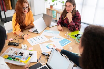 High angle of crop anonymous female office workers sitting at table with laptop and paper charts while discussing and working on project together in office