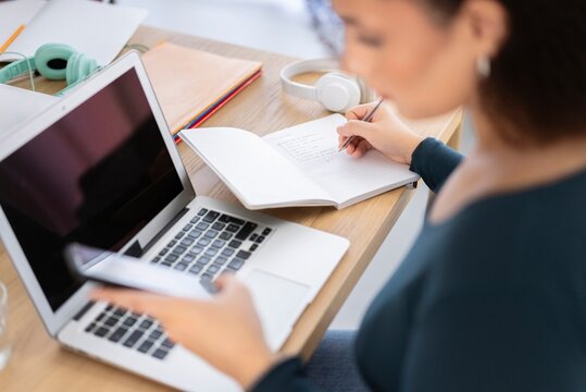 Side view of crop anonymous female worker sitting at table with laptop and browsing smartphone while writing on notebook and working on project in office