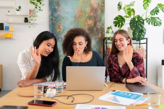 Group of happy young coworkers sitting at table with laptop while waving hands during video call meeting with colleague in modern workspace at daylight