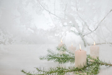 Four lit white candles and a fir tree branch in front of frozen glass and a snowy winter landscape,...