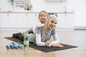 Adorable girl in sport outfit lying on mothers back with positive energy during exercises. Loving muslim female in hijab relaxing with cute daughter after fitness routine at home.