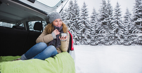Woman in woolen hat sits in the trunk of the car and holds a cup of hot tea in her hands - obrazy, fototapety, plakaty