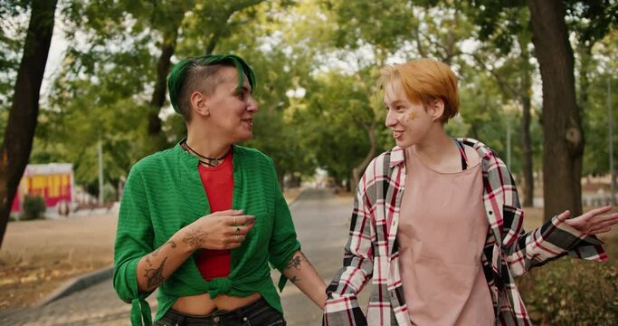 Front view of two lesbian girls with bright short hair in shirts walk through the park during their date and communicate