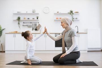 Muslim mother and little daughter engaged in fitness, yoga, exercise at home. Smiling woman in hijab and small kid girl sitting on the carpet floor on knees and giving high five looking at camera.