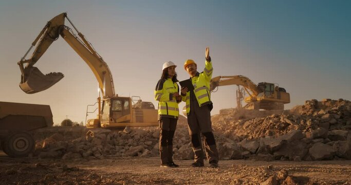 Hispanic Female Inspector Talking to Caucasian Male Land Development Manager With Tablet On Construction Site Of Real Estate Project. Excavators Preparing For Laying Building Foundation. Hot Sunny Day