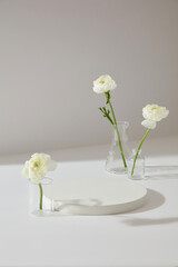 Minimal scene of white background featured few Persian Buttercup flowers inside glassware. Blank round podium to display your cosmetic product or goods