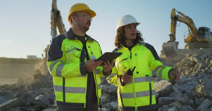 Caucasian Male Civil Engineer Talking To Hispanic Female Architect And Using Tablet On Construction Site of New Apartment Building. Real Estate Developers Discussing Business, Excavators Working.