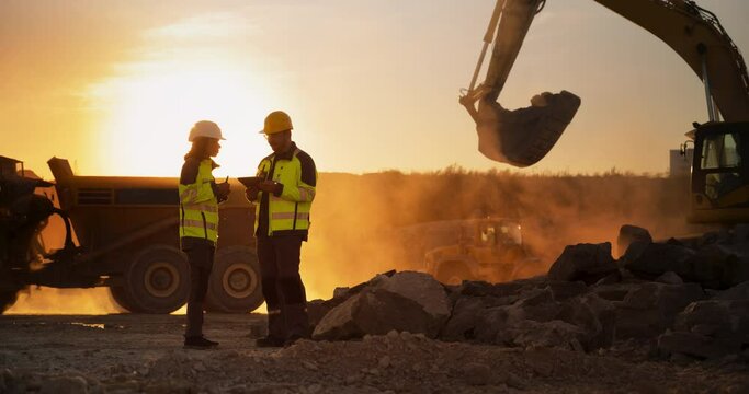 Cinematic Golden Hour Shot Of Construction Site: Caucasian Male Civil Engineer And Hispanic Female Architect Talking And Using Tablet. Trucks, Excavators, Loaders Working To Build New Apartment Block.