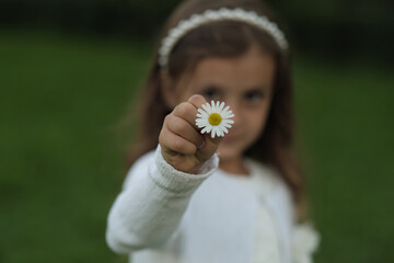 Portrait of a young girl in a white dress holding a daisy close to the camera, with a soft focus green background.