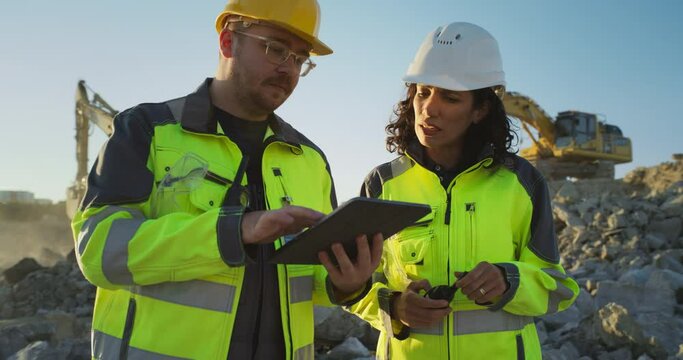 Caucasian Male Civil Engineer Talking To Hispanic Female Inspector And Using Tablet On Construction Site of New Apartment Building. Real Estate Developers Discussing Business, Excavators Working.