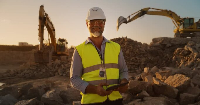 Successful Bearded Civil Engineer Wearing Protective Goggles And Smiling At Camera On Construction Site on a Sunny Day While Heavy Machinery Working. Man Holding Tablet, Wearing Hard Hat, Safety Vest.