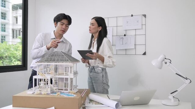 Beautiful young caucasian project manager brief asian engineer by using tablet while engineer measuring house model on meeting table with blueprint, architectural equipment scatter around. Immaculate