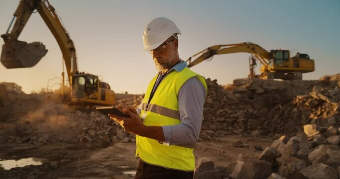 Caucasian Male Civil Engineer Wearing Protective Goggles And Using Tablet On Construction Site On A Sunny Day. Man Inspecting Building Progress. Excavator Loading Materials Into Industrial Truck.