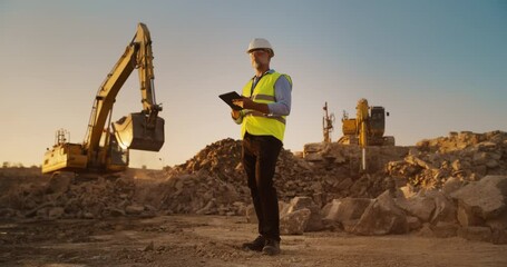 Caucasian Male Real Estate Investor Wearing Protective Goggles And Using Tablet On Construction Site On A Sunny Day. Man Inspecting Building Progress. Excavator Loading Materials Into Industrial Truck - Powered by Adobe