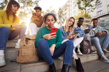Diverse group of university friends sitting on street staircase, serious students people using and looking at smart phones. Photo shows addiction to new technologies and social networks, generation Z. - obrazy, fototapety, plakaty
