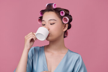 Happy young woman in dressing-gown and hair curlers drinking coffee on pink background