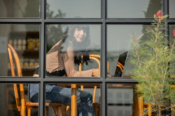 Positive emotions. Lifestyle concept. Charming asian woman smiling, looking camera with relaxed expression sitting inside coffee shop viewed through glass with reflections as they sit at a table.
