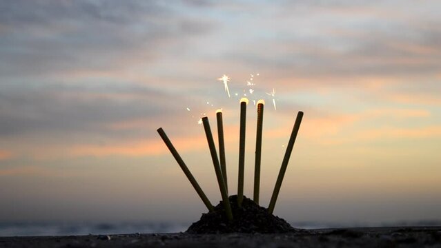 Fire fountain candles burning sparkling on the background sky during sunset at dawn in sand beach of seashore. Festive fireworks holiday. Glowing Bengal light. Concept celebration New Year Christmas