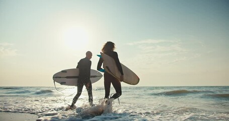 Two surfer guys in wetsuits walk into the water holding their surfboards on a sunny summer morning. Two guys surfers on the beach in summer