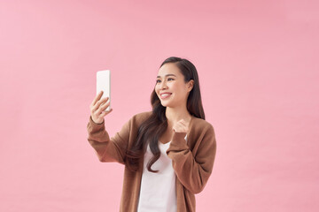 Pretty young woman with smartphone taking selfie against pink background