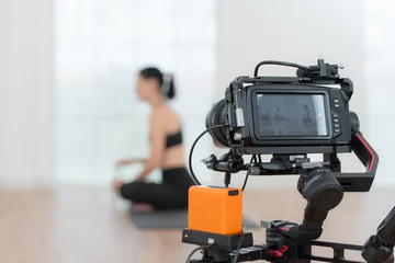 Abwaschbare Fototapete Blogger recording live video tutorial at home. Young woman sitting on yoga mat in lotus position and holding a camera. © Wosunan