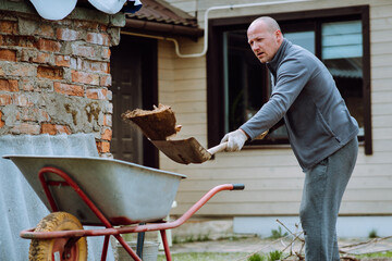 Mature man with bald head throwing earth into wheelbarrow using shovel. Gardener in gloves collecting land on yard.