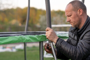 Middle aged human with furrowed eyebrows carefully fastening metal posts to trampoline support outdoor using bolt.