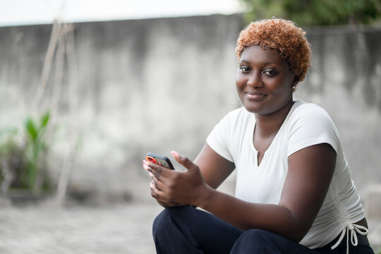 image of beautiful blonde african lady with smart phone outside- cheerful young black model seated outdoor