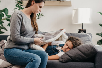 Caring young caucasian mother covers blanket sick preteen daughter lying on couch in living room at...