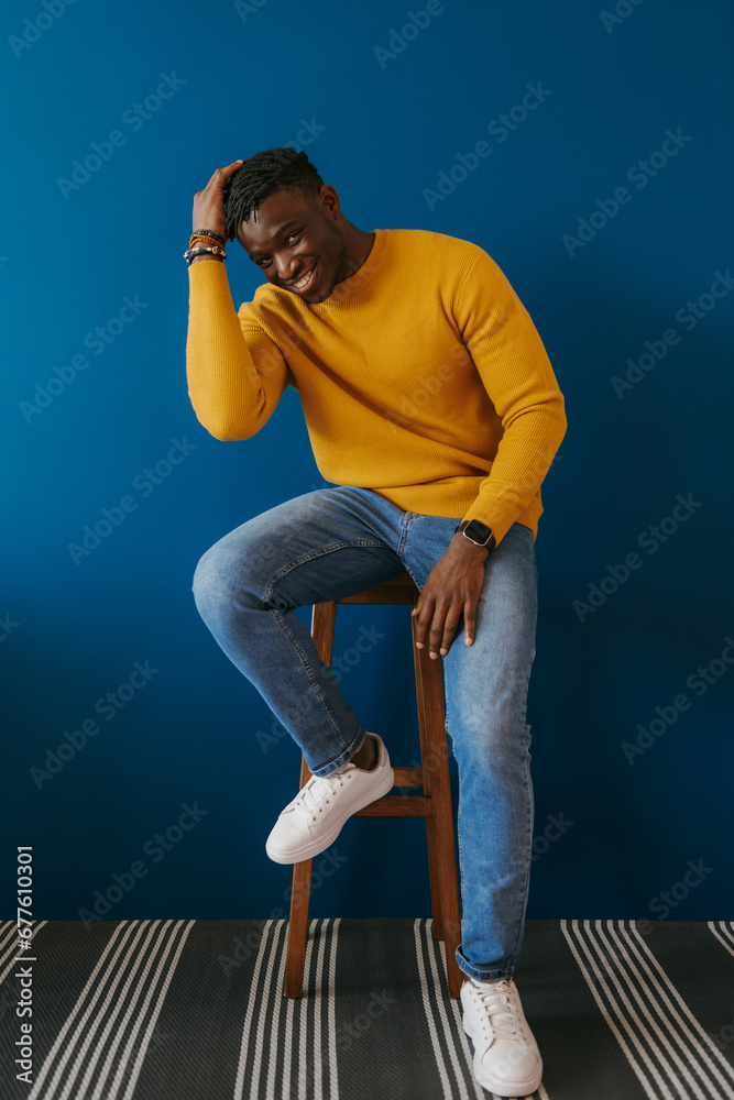 Wall mural Charming young African man in casual wear looking relaxed while sitting on the chair against 