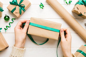 Woman is packing surprise gift for Christmas holidays, sitting at workplace with craft tools, top view. Woman wrapping Christmas gift at colored table
