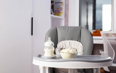 A baby bottle with milk and a plate with dry milk formula enriched with bifidobacteria, vitamins and minerals stands on a gray high chair in the children's room.