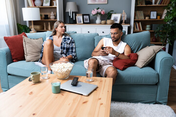 Young boyfriend making fun and jealous his girlfriend pretending that he is texting with other girl on his cell or mobile phone while they sitting on the sofa in their apartment - obrazy, fototapety, plakaty