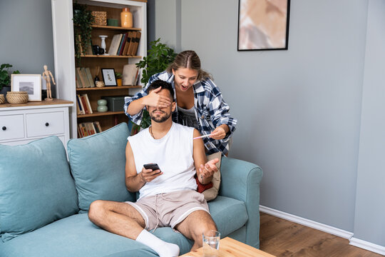 Happy wife makes surprise to husband, covers his eyes and holds pregnancy test, informs he will become father going to share good news, pose together in bedroom. Joyful future parents